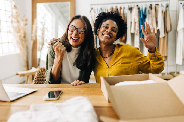 Two women in a small business workspace celebrating with laptops and boxes, embodying teamwork and success.