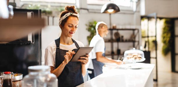 Smiling small business owner checks inventory on tablet, with employee preparing products in the background.